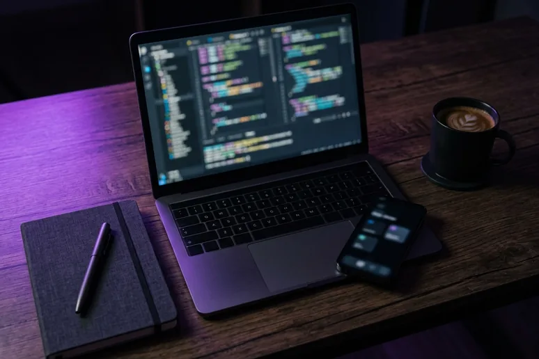 Escritorio de desarrollo visto desde arriba de noche: portátil abierto con código desenfocado en pantalla, móvil, libreta y bolígrafo, y una taza de café sobre mesa de madera oscura con luz morada