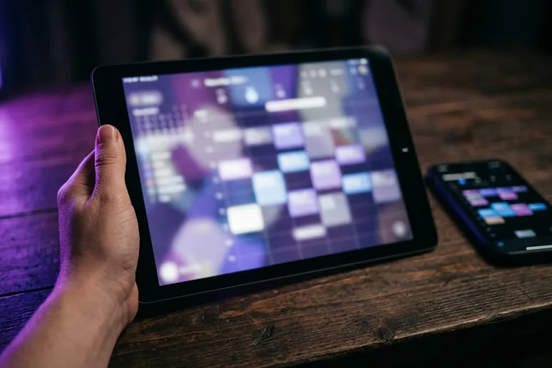 Una mano sostiene una tableta con una interfaz de calendario desenfocada, con un móvil al lado sobre una mesa de madera y luz de acento morada